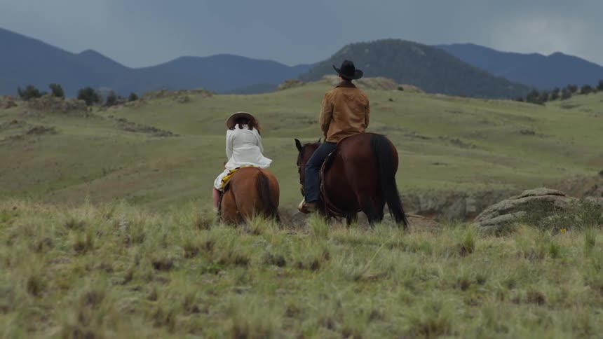 A man and a woman are riding horses forward in a grass field with some hills in the background