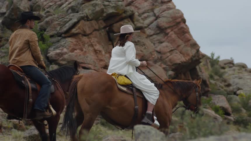 Man and woman riding horses through a field stop near a rock formation on an overcast day