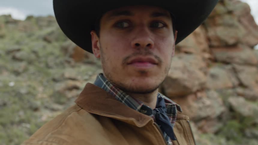 A man wearing a cowboy hat looks around while standing near a rock formation