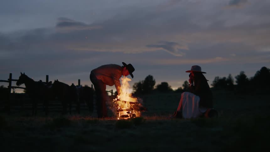 A man tending to a campfire while a woman sits nearby at dusk