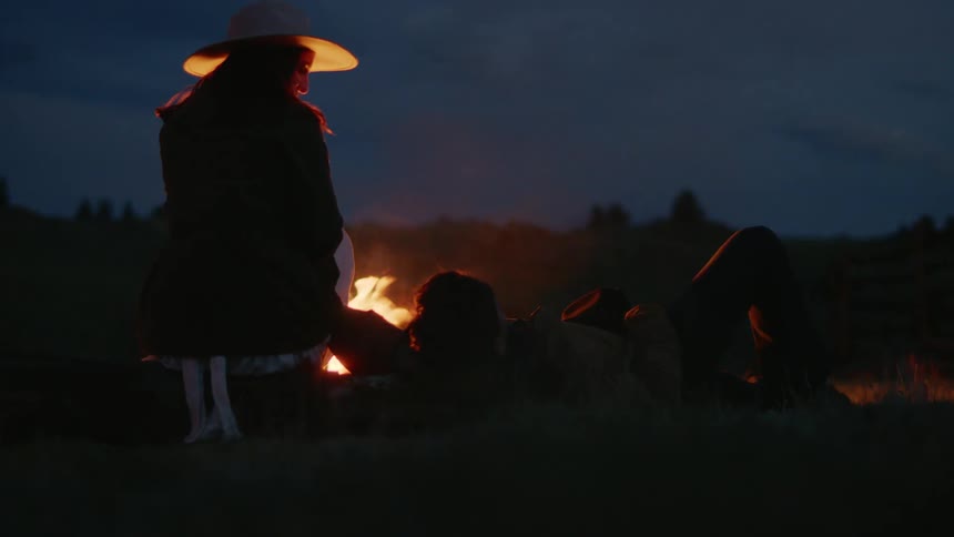 Woman sits near campfire at night while her male friend lies on his back