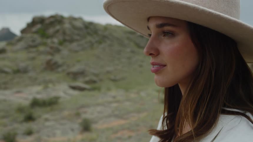 A woman with long hair wearing a wide brimmed hat a rocky natural landscape in the background