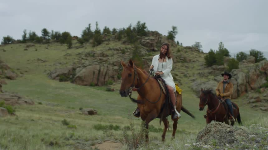 A man and a woman riding horses in a grass field with hills and the woman is smiling