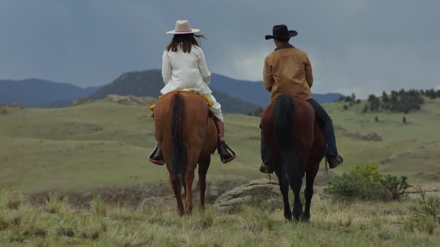 A man and a woman ride horses with the woman stopping to take a picture of a large grassy area and mountains