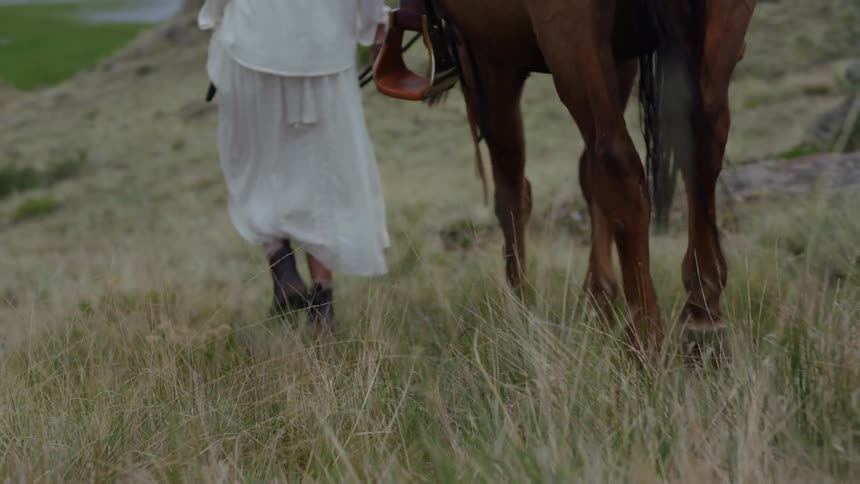Man and woman walk down a grassy slope toward the river leading their horses