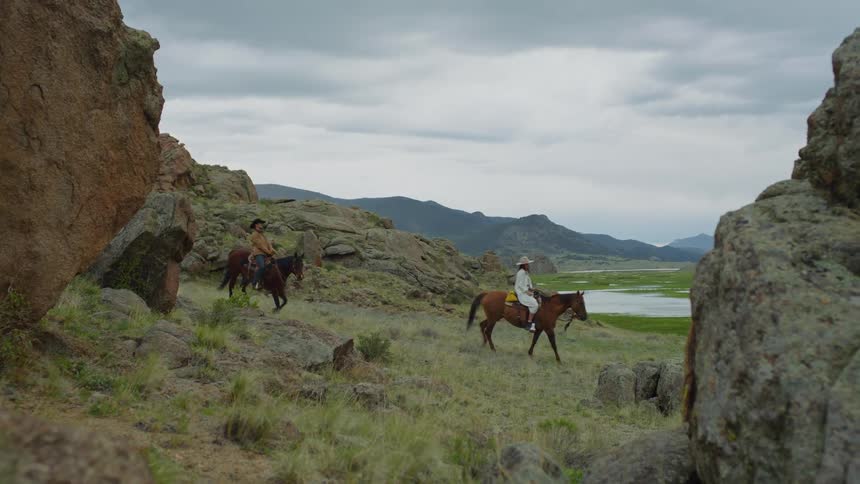 Man and woman riding horses in a rocky environment with a lake and mountains in the background