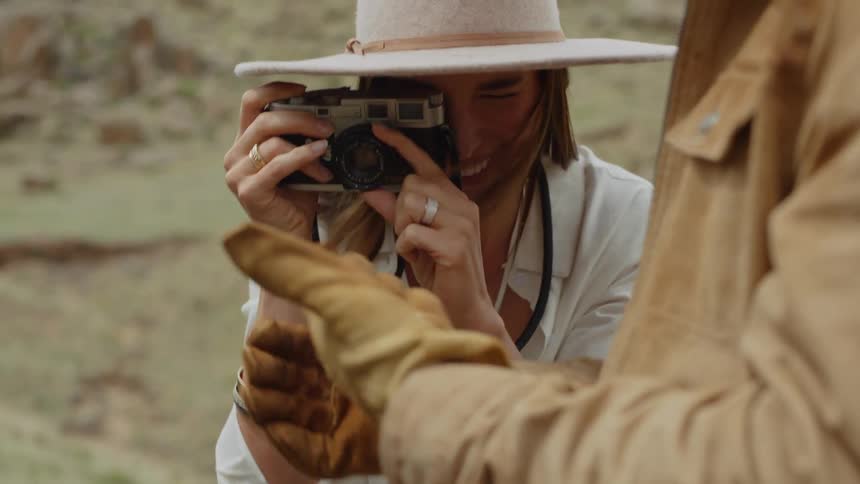Standing near a hill a smiling female photographer takes pictures of a bug held by a man wearing a cowboy hat