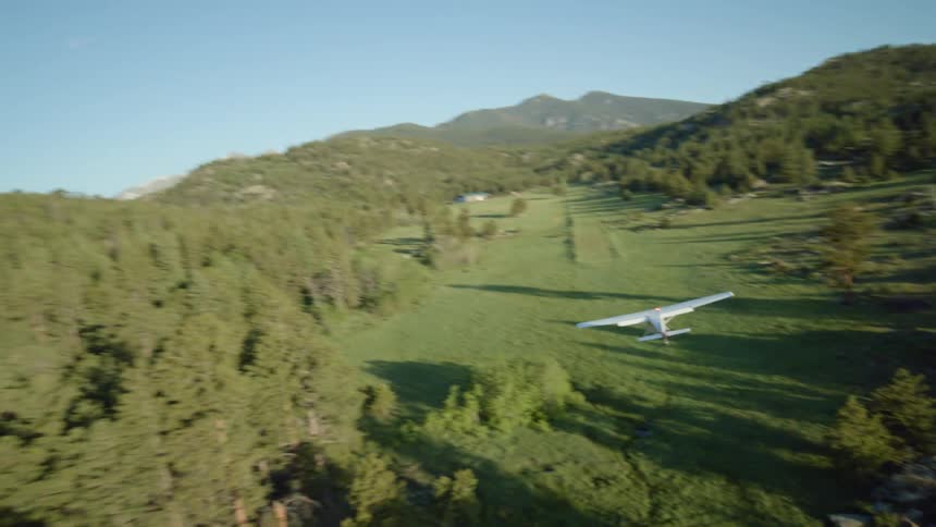 A small airplane lands in a field near the forest in front of the mountains
