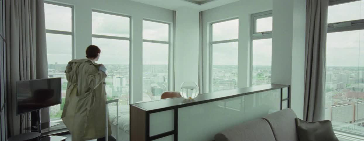 A person arrives in a room with several large glass windows and he takes off a coat and goes to look at the view through the window