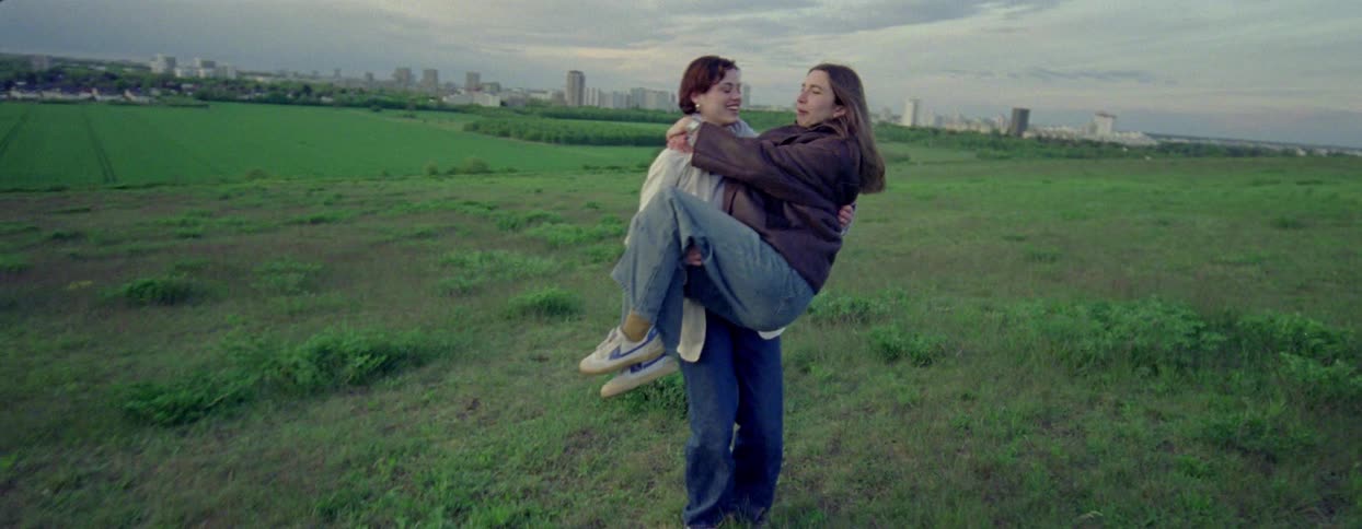 Two women laugh as one of them carries the other in a field outside the city on an overcast day
