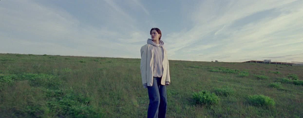 A young woman is standing with her back turned and walking on a flat field with grass and then she looks back