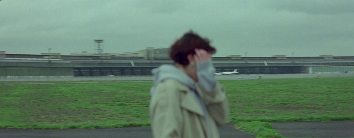 Female traveler walks along the road with her luggage across a grassy field from the airport on an overcast day