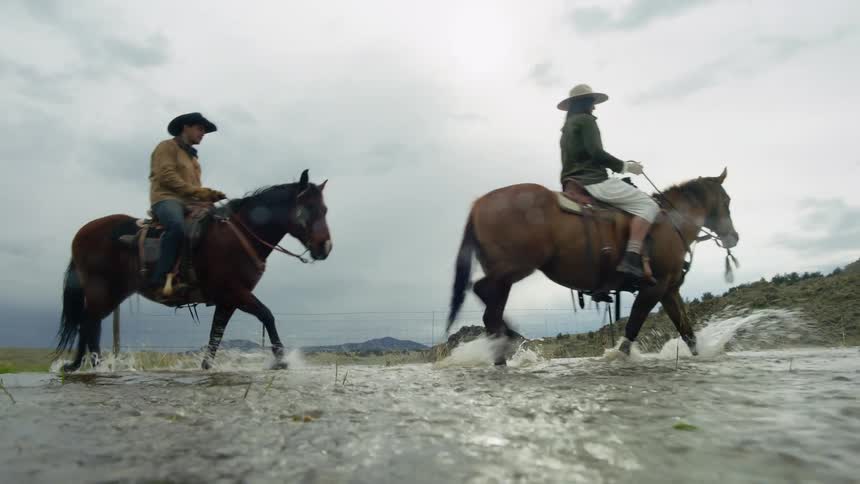 Two individuals riding horses through shallow water under overcast skies