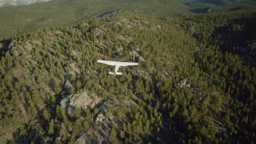 A small white plane flying over a region with hills full of pine forests
