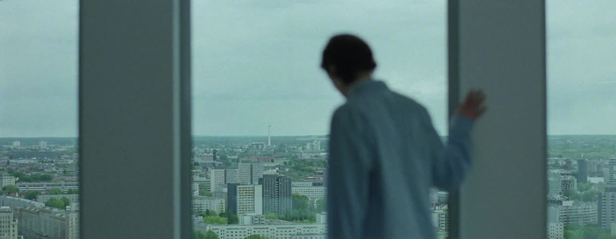 Woman walks back and forth stopping to look out the window at the city on an overcast day