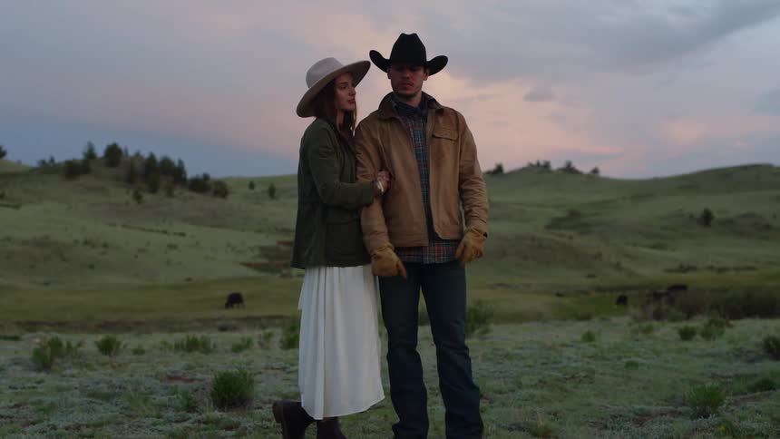 Woman hugging a man both wearing hats in a field with hills in the background on a cloudy late afternoon