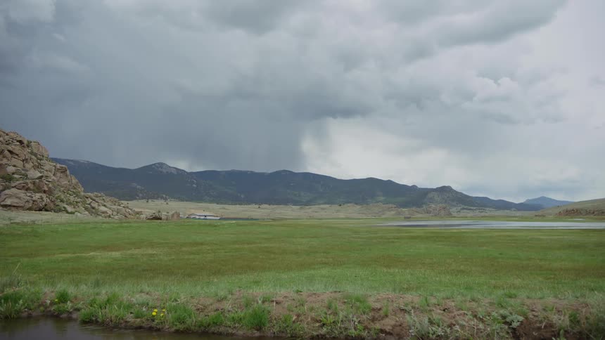 A landscape of a huge field of green grass with a river running in front and in the background is a mountain range and the sky is with gray clouds