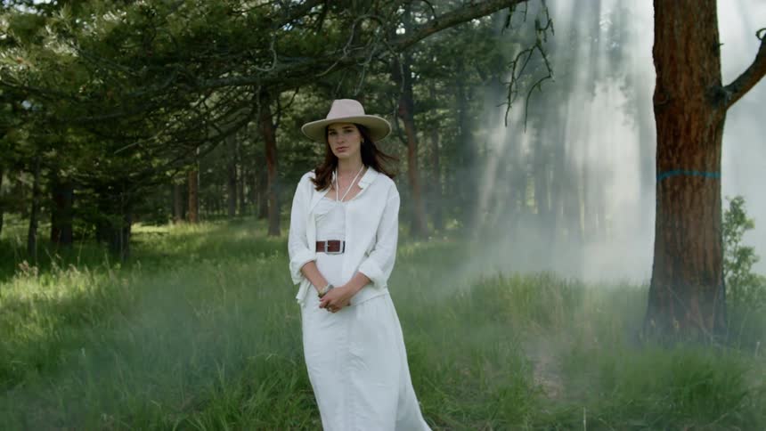 Woman wearing a hat and a long white dress in the middle of a smoky swamp