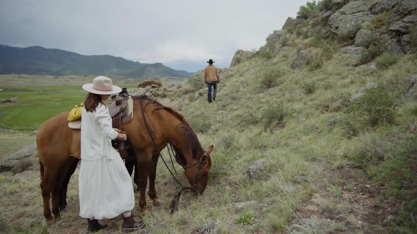 Woman adjusts the saddle on a horse while a man walks on a mountain