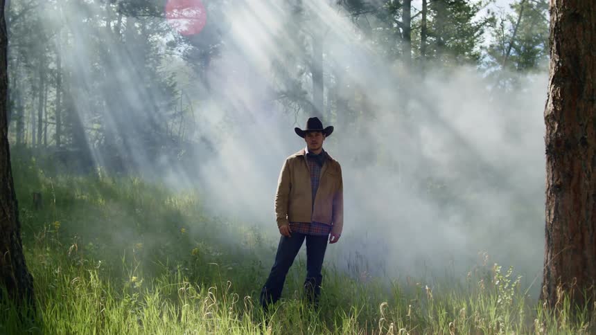 A cowboy stands in a forest as a fire pumps smoke into the sky