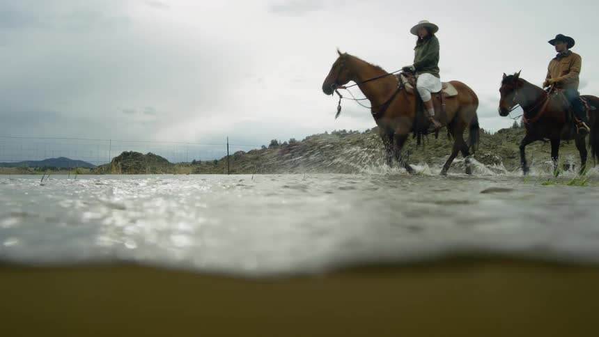 Two people ride their horses through the water