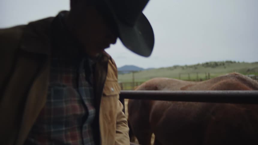A cowboy carries a bale of hay in a barn next to a horse corral