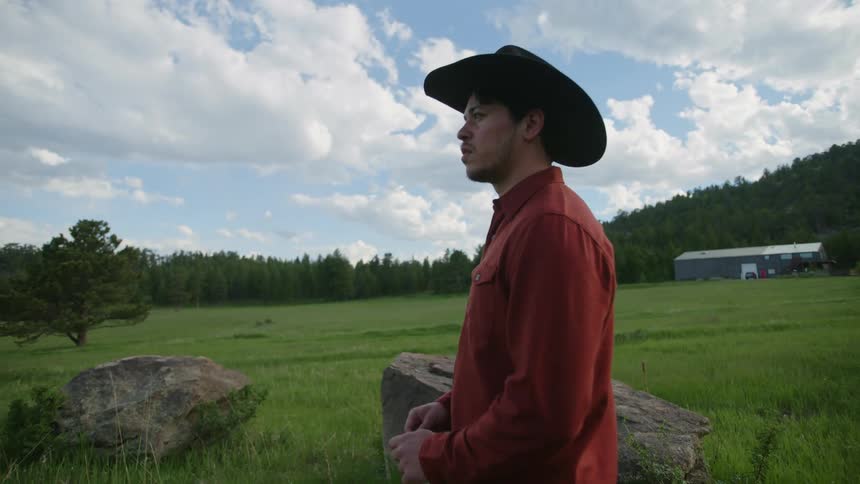 A cowboy standing in the green grass looking sad turns and walks away