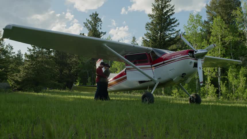 Couple saying goodbye in a green field with trees around and the woman getting into a small plane