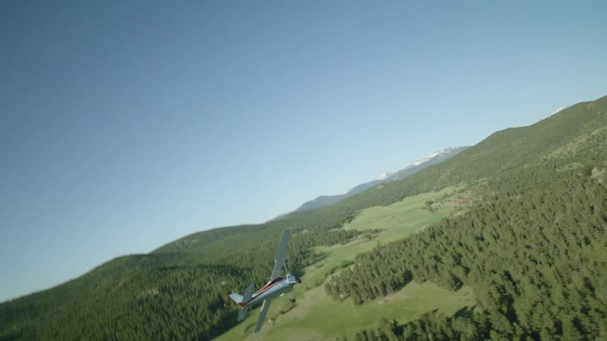 Aerial view of a small private jet flying above mountains and forests on a clear blue sky day