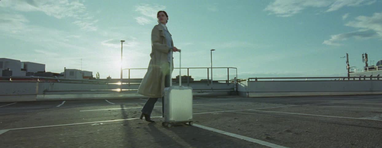 A woman in a trench coat walking with a suitcase in an empty parking lot under a blue sky