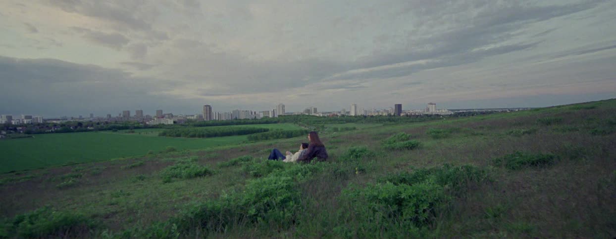 Two women lying in a vast field with a metropolis on the horizon on a cloudy day