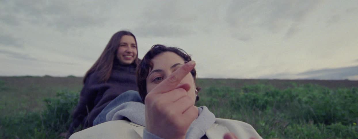 Two women wearing jackets recline in a field and laugh while discussing an insect
