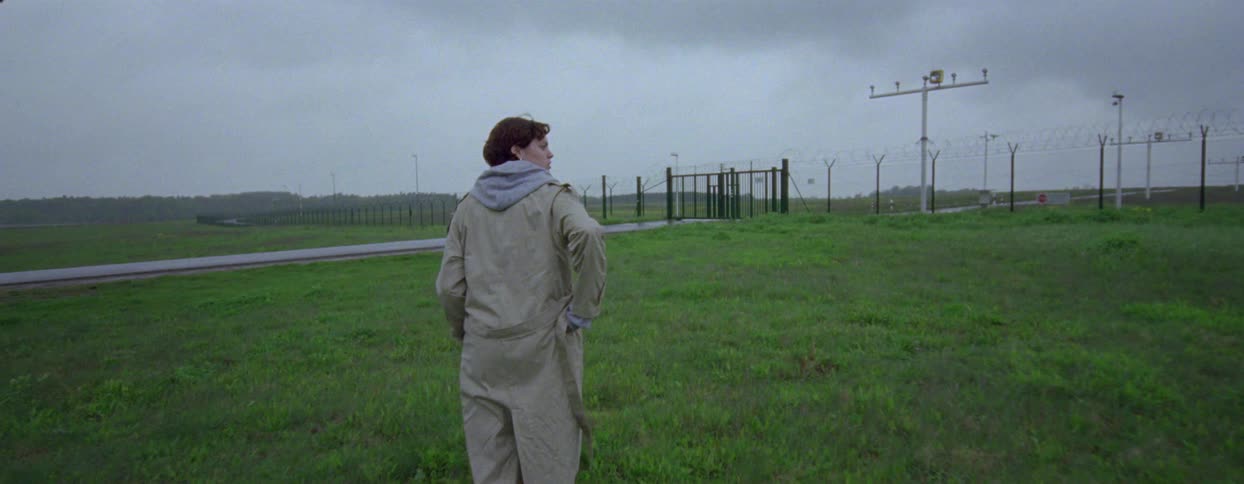 A woman walking in a grassy field under overcast skies with a fenced area and security features visible in the background