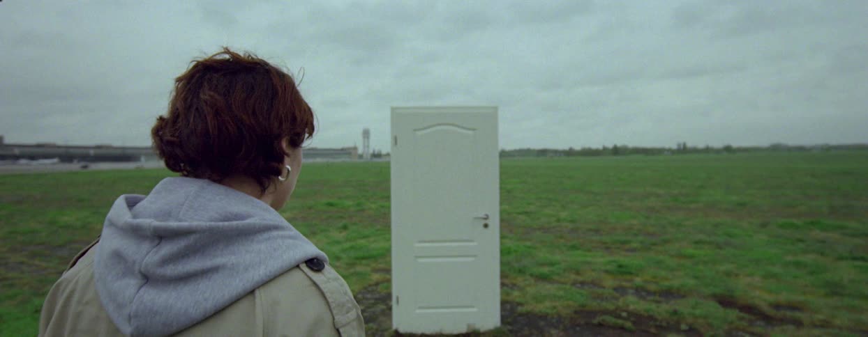 A woman looks creeped out at a door standing in the middle of a large grassy field