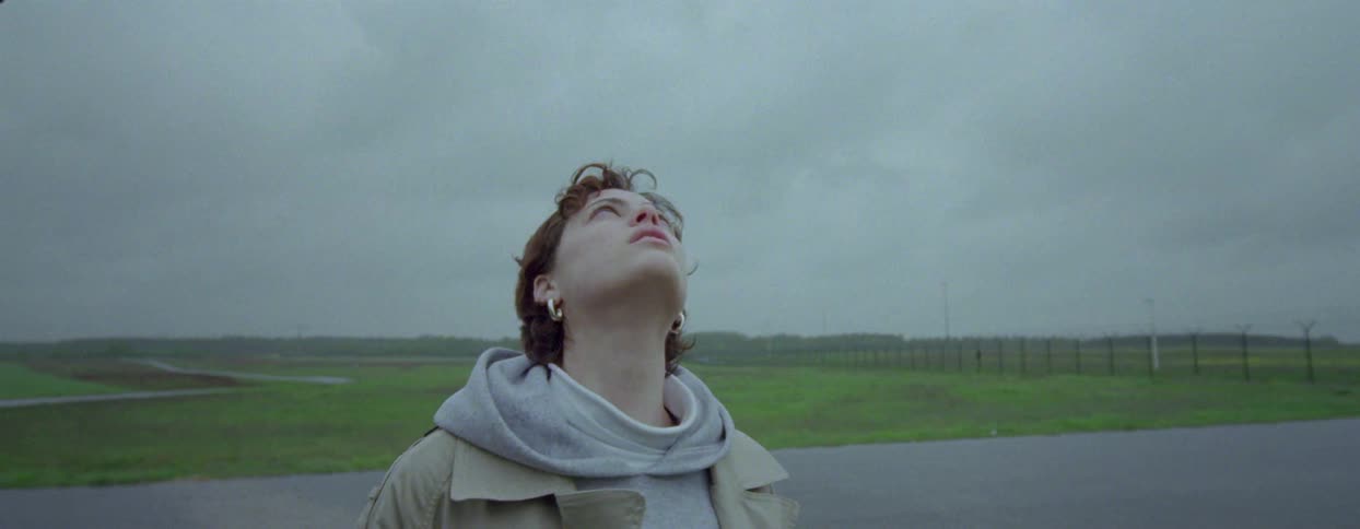 A young woman is looking at a plane that is passing in the gray sky and she is following it with her eyes