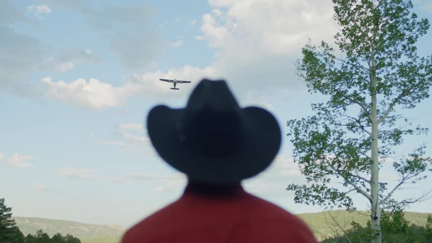 A man wearing a cowboy hat watches a plane fly by overhead