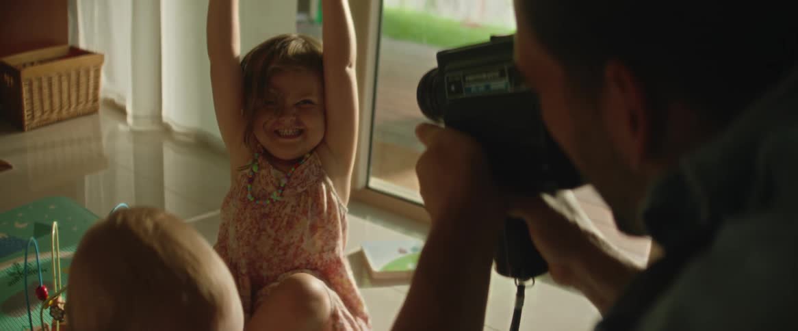 A father filming with a camera as his daughter lifts up a book with a smile