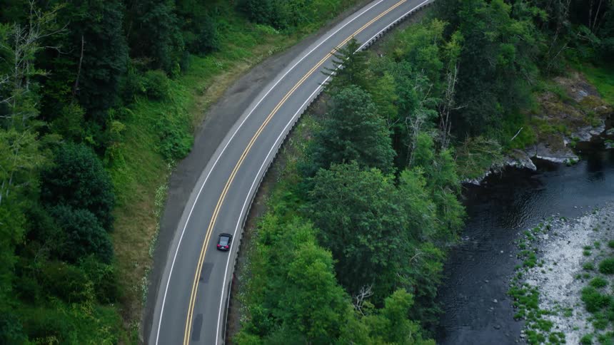 Car drives through the forest along a highway curving around a river during the day