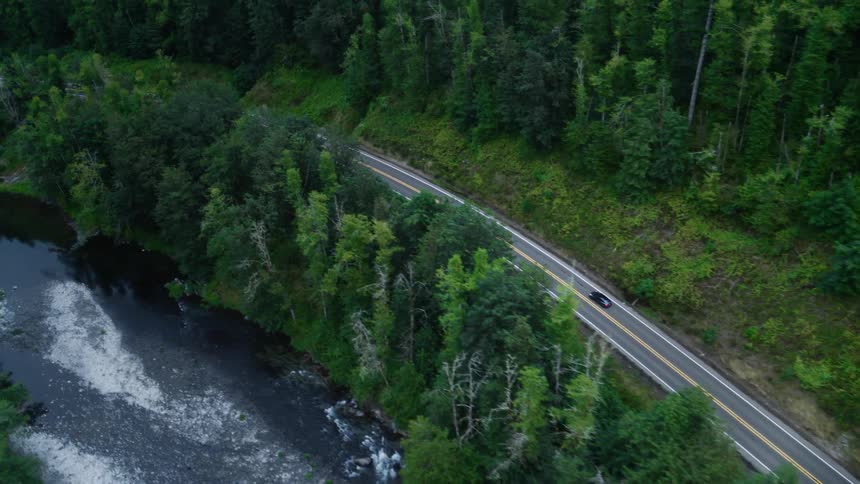 Aerial view of a vehicle traveling on a highway surrounded by dense forests and the edge of a river