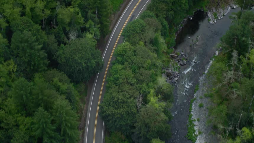 An aerial view shows a car driving on a road through a forested area with low clouds overhead