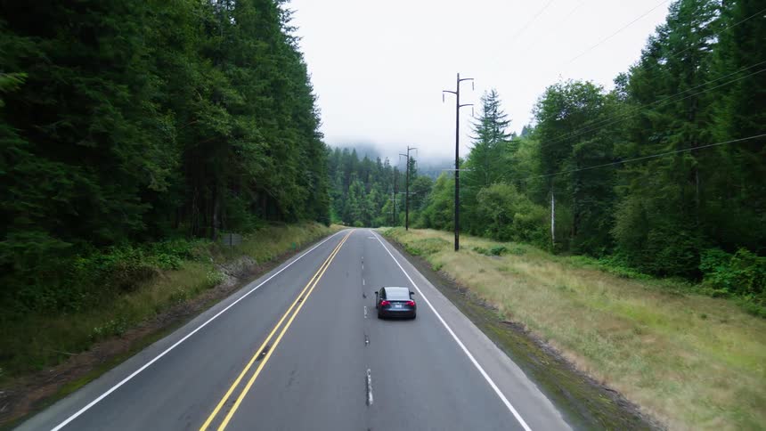 An aerial view shows a car driving on a road through a forested area with low clouds overhead