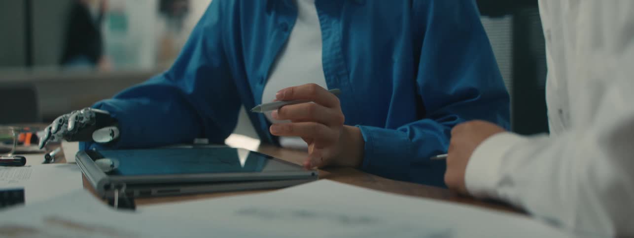 Arms of two people sitting at a table with one of them having a robotic hand holding a pen and using a tablet