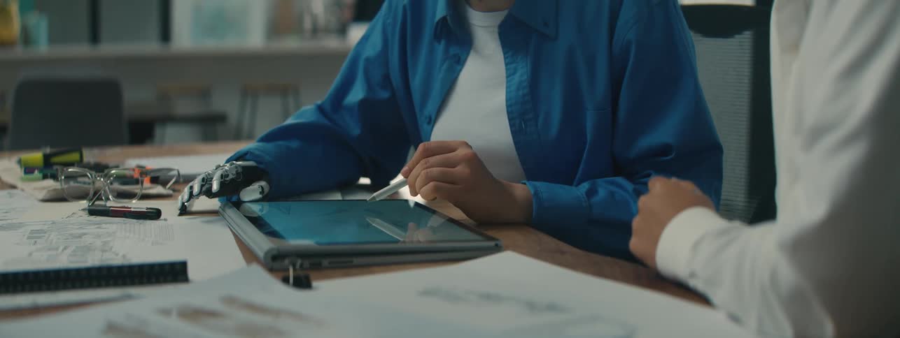 A woman with a prosthesis for her arm uses a tablet and stylus to explain something to another person
