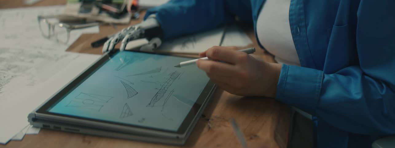 A woman sits at a table and draws designs on a laptop