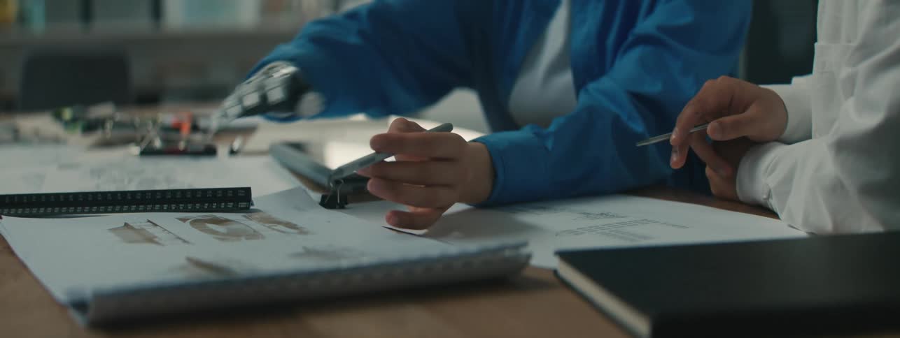 Person with a robotic hand holding a pen and explaining content with books around to a colleague next to him