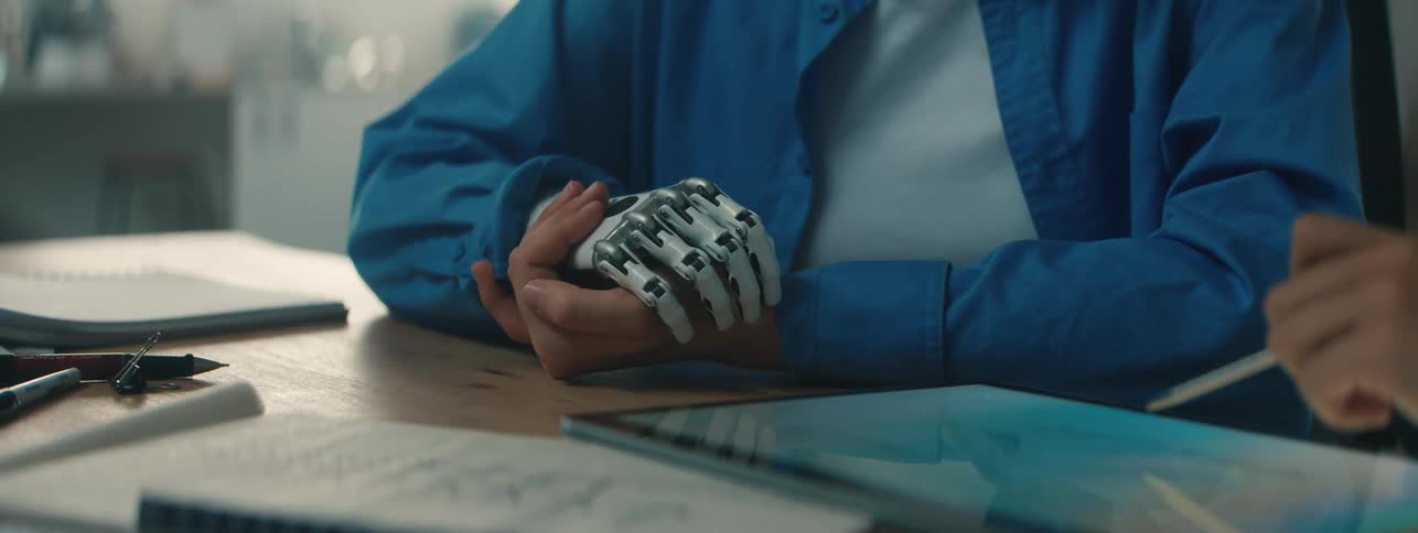A healthcare worker writes on a tablet while a patient with a prosthetic hand sits nearby