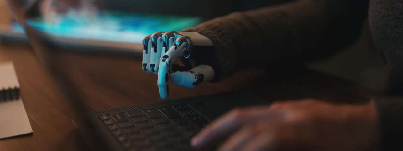 A person with a robotic prosthesis hand with metal components presses keys on a laptop keyboard