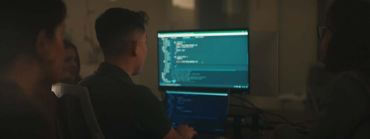 Group of people sitting at a table facing a computer in a dimly lit room