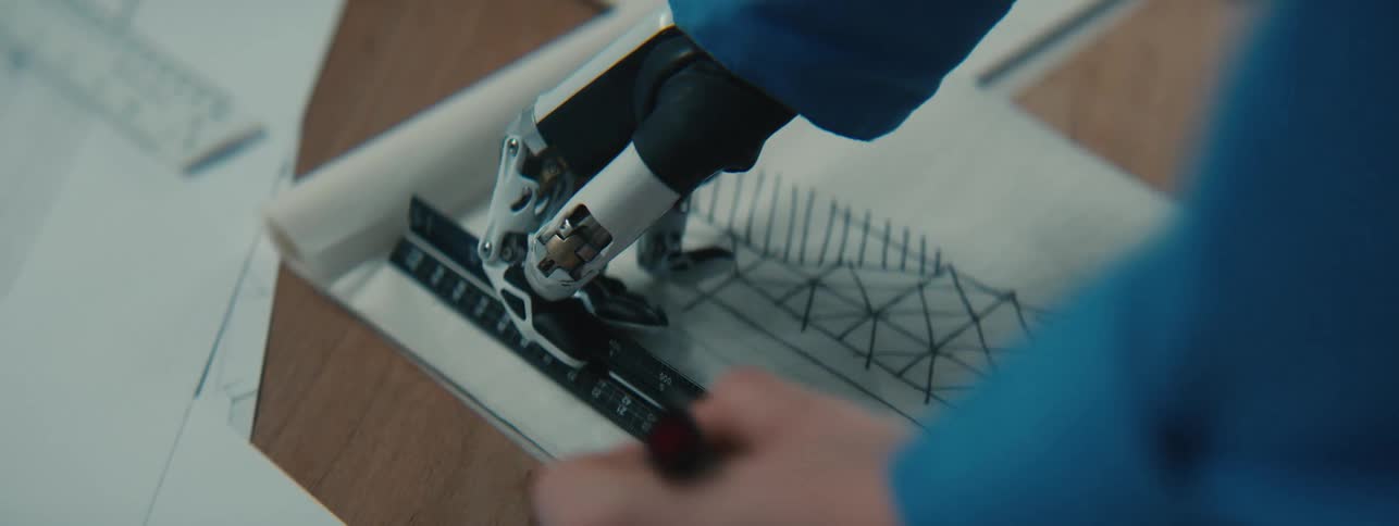 A woman uses her mechanical prosthetic hand to hold a ruler in place over a paper while she uses it to draw a straight line with her other hand