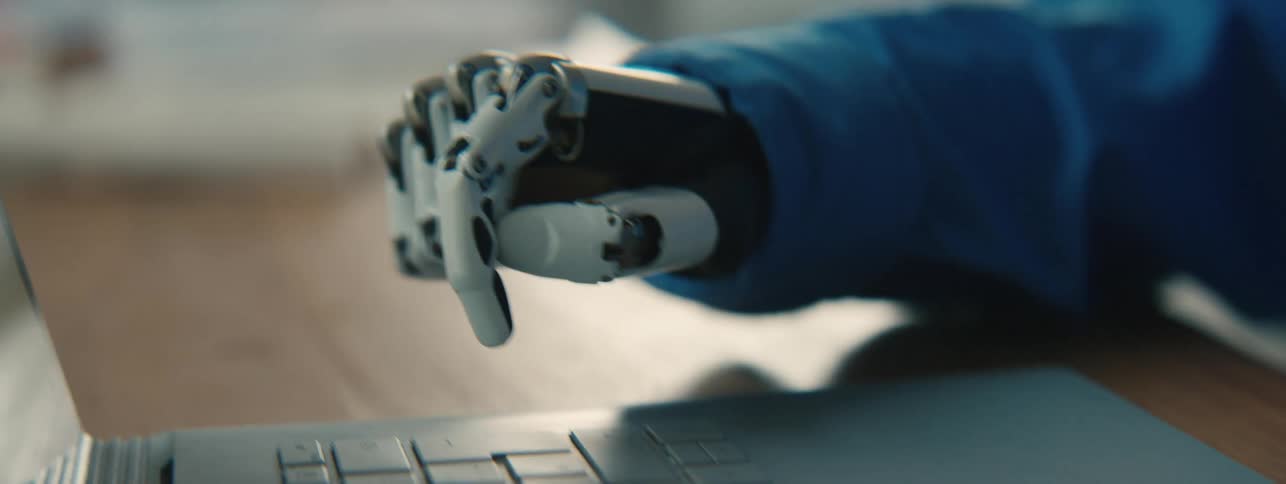 A mechanical prosthetic hand typing slowly on a laptop keyboard